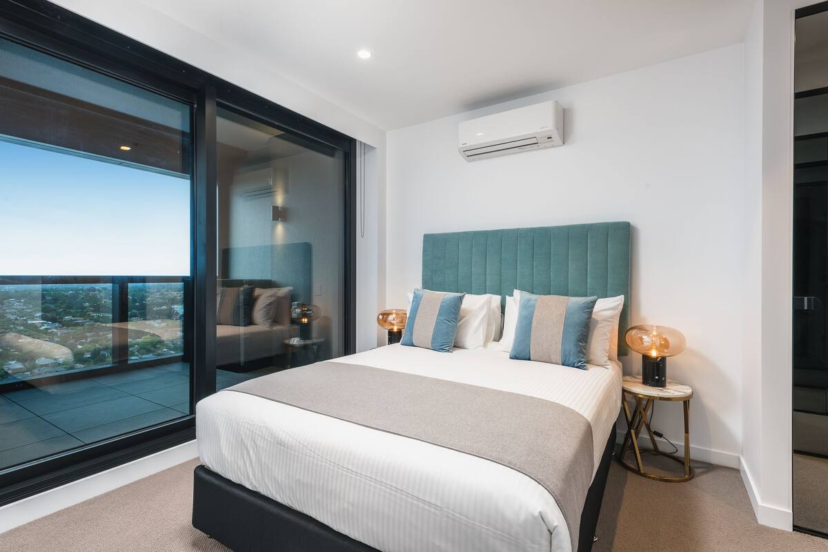 A modern bedroom with a large bed, a view through a sliding glass door, and a small bedside table with a lamp.