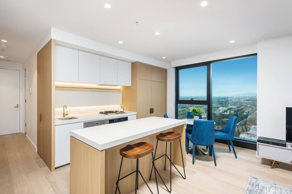 A modern kitchen with a white island, wooden accents, and a large window offering a city view.