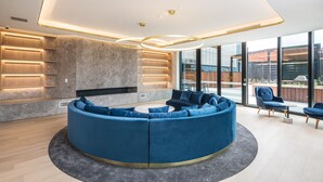 A modern lounge area with a curved blue sofa, wooden shelving, and a large window offering a view of the outdoors. - Sky One Apartments by CLLIX (Box Hill)