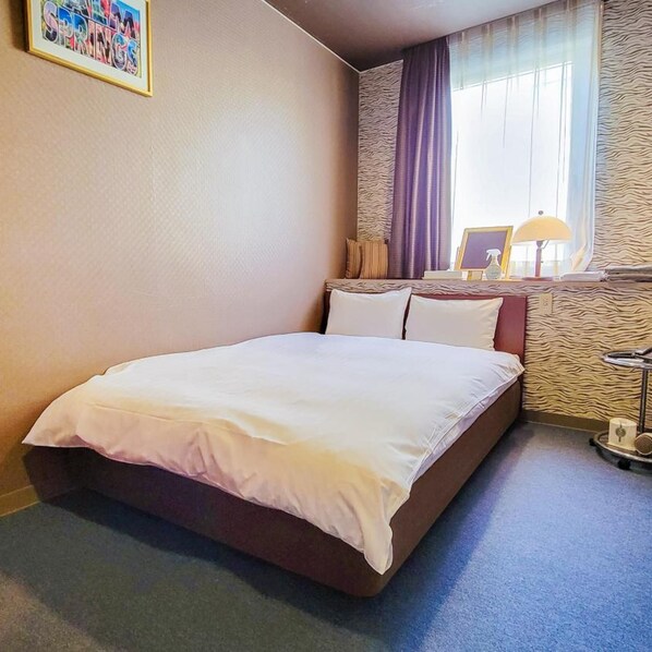 A hotel room with a bed, a window with curtains, a wall-mounted lamp, and a framed picture on the wall. - Hotel Palms Tenmonkan (Kagoshima)