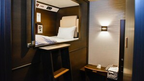 A compact hotel room with a single bed, a small table, a chair, a bedside table, and a wall-mounted light fixture. - Hotel Palms Tenmonkan (Kagoshima)