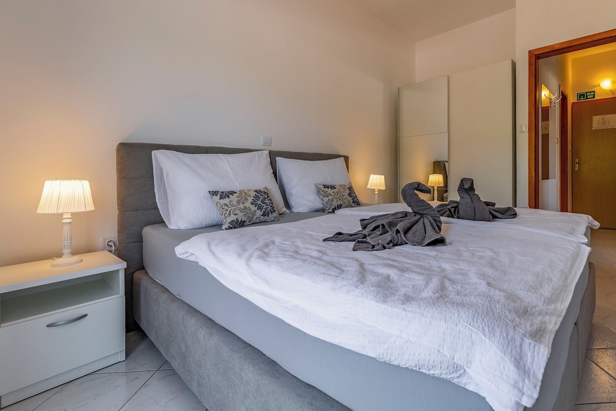 Ein ordentlich bezogenes Bett mit weißer Decke, grauen Kissen und zwei Deko-Schwanen aus Handtüchern. Links befindet sich eine Nachttischlampe und rechts eine Tür mit einem Ausgangsschild.