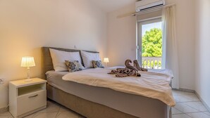 A neatly made bed with a blanket and two decorative pillows, a bedside table with a lamp, and a view of greenery through a window. - Apartmani Rok (Medulin)