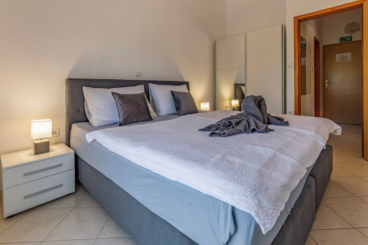 Ein ordentlich bezogenes Bett mit weißer Decke, grauer Decke und einem aufgebogenen Handtuch. Ein Nachttisch mit Lampe, Schublade und kleinem Schrank. Das Zimmer hat eine Holztür und einen Fliesenboden.