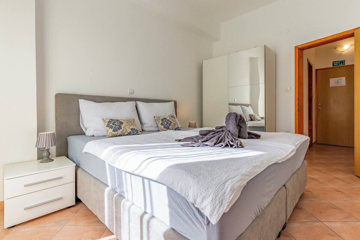 Ein ordentlich bezogenes Bett mit grauer Decke und Kissen, ein Nachttisch mit Lampe und eine Holztür rechts.