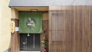 A building entrance with a wooden door, a green banner with Japanese characters, and a potted plant. - Shirakabanoyado-Uehonmachi (Osaka)