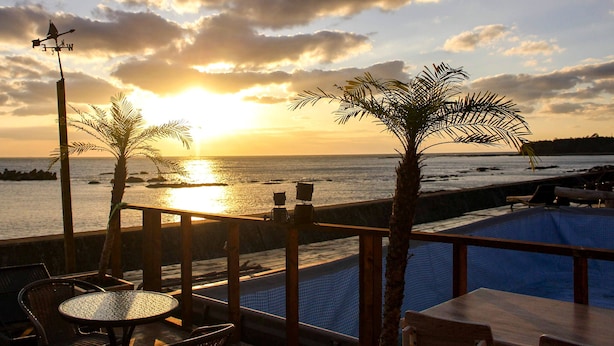 Zona junto a la piscina con vista al atardecer sobre el océano, palmeras y un veleta.