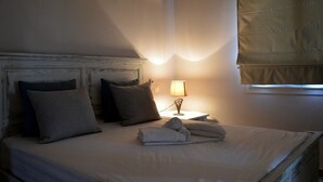 A neatly made bed with pillows, a bedside lamp, and a window with blinds. - Ammos Luxury Suites (Monemvasia)