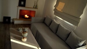 A modern living room with a grey sofa, a fireplace, and a window with blinds. - Ammos Luxury Suites (Monemvasia)