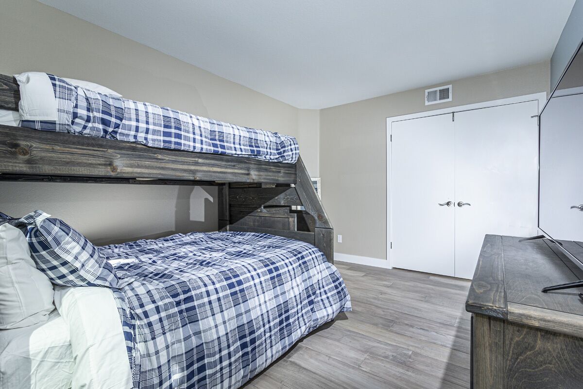 A bunk bed with a checkered bedspread, a wooden desk, and a white wardrobe in a room with wooden flooring.