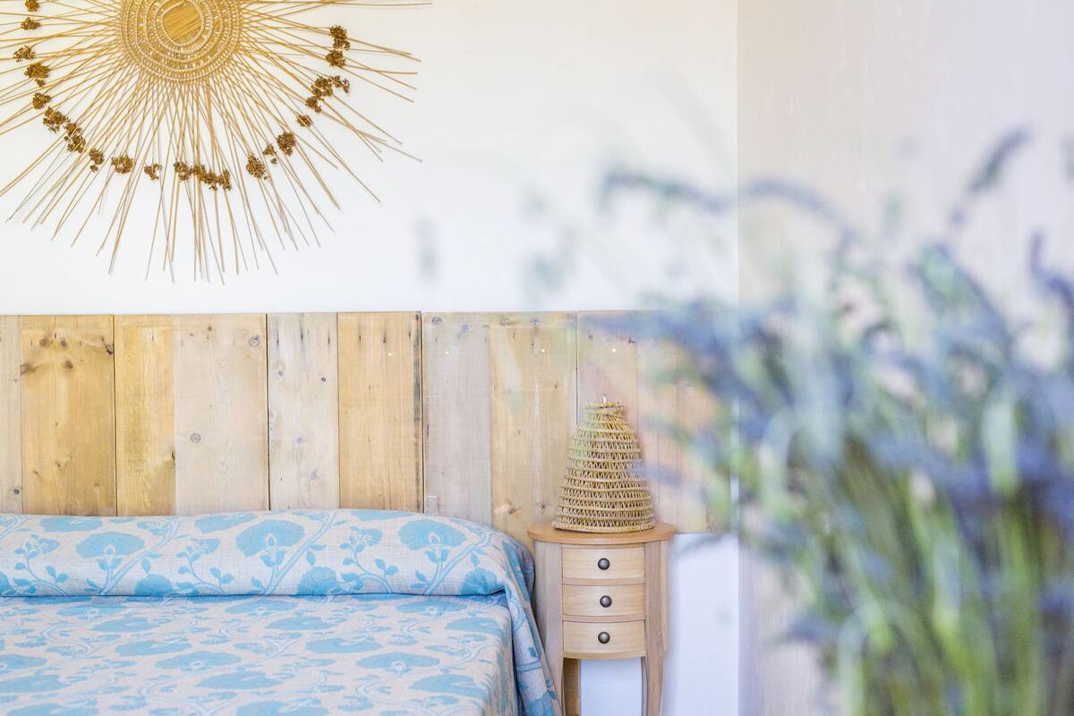 Een slaapkamer met een houten hoofdbord, een bed met een blauw beddegoed met patroon, een nachtkastje met een geweven lamp en een wanddecoratie.