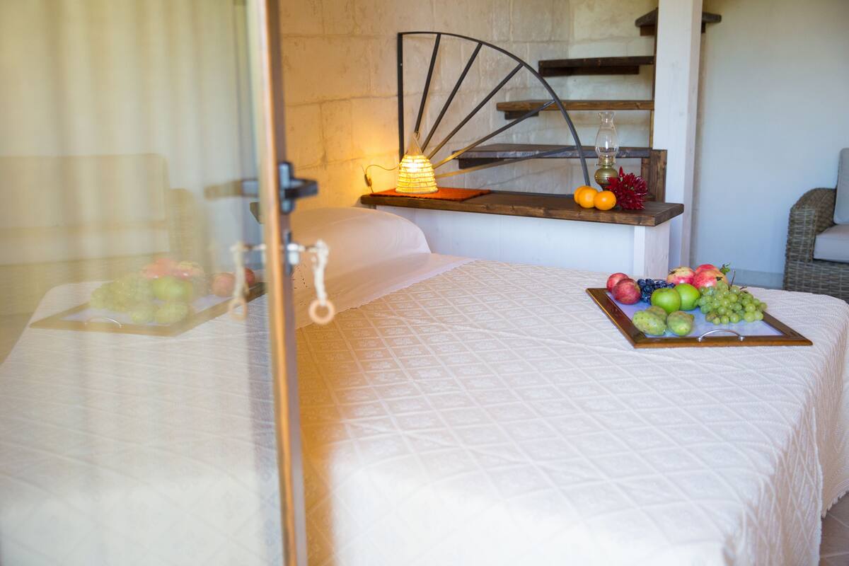 Een keurig opgemaakt bed met een schaal fruit en een klein tafeltje met een lamp en sinaasappels.