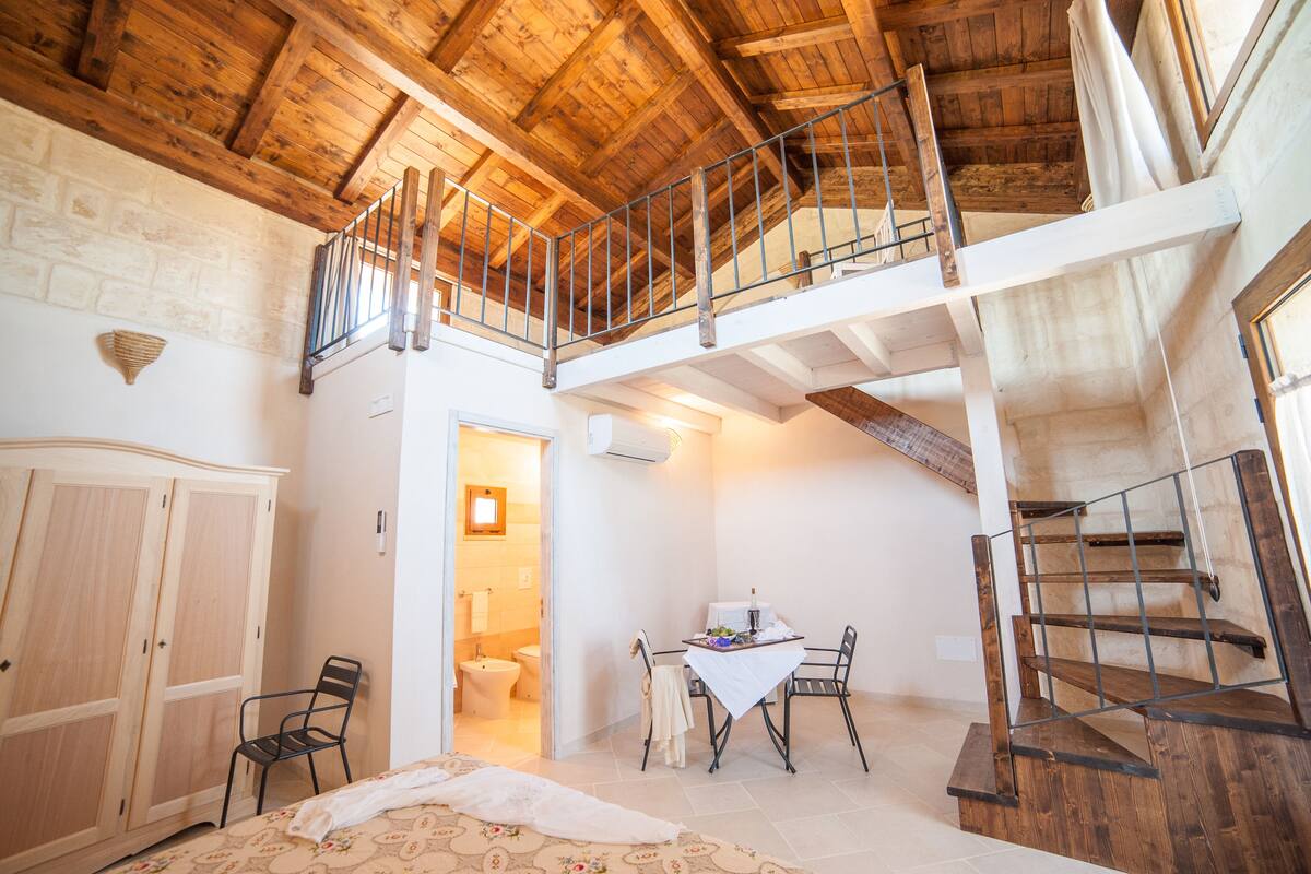 Een ruime kamer met houten plafond, een balkon, een eethoek met tafel en stoelen, en een trap.
