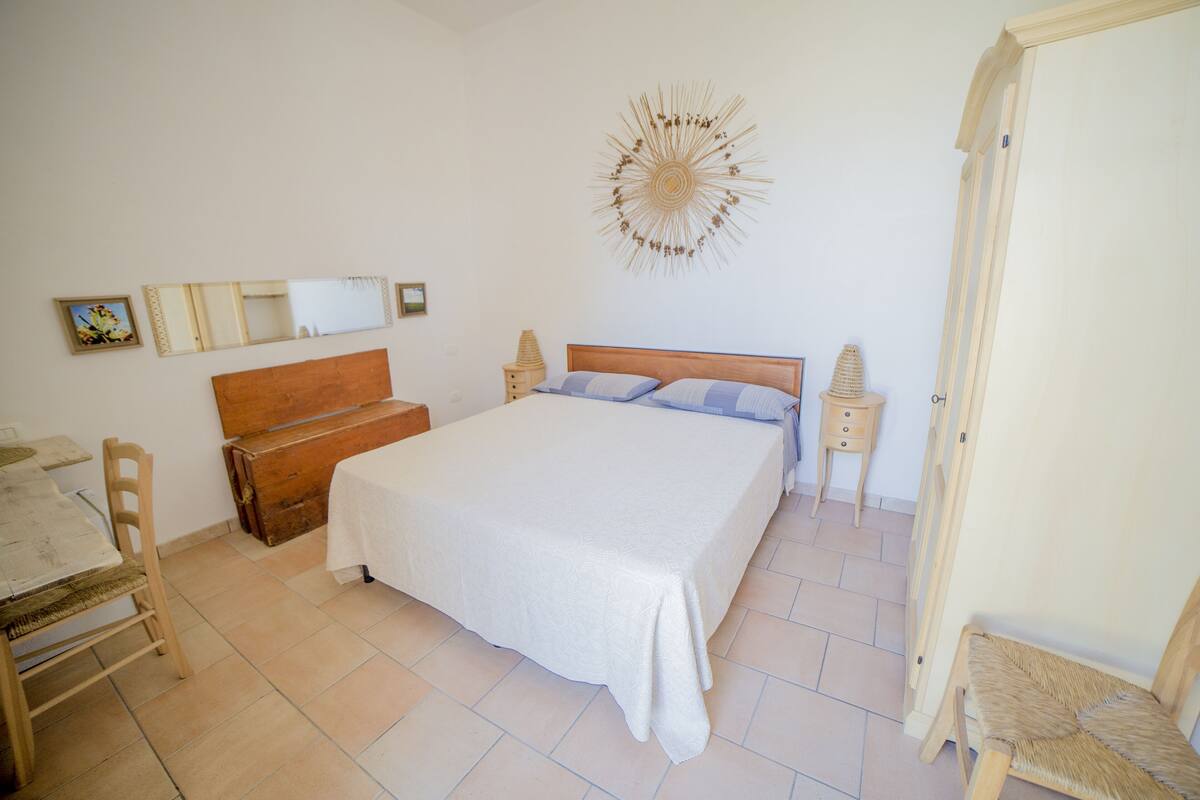 Een slaapkamer met een groot bed, een houten kist, een stoel, een klein tafeltje en een spiegel aan de muur.