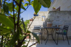 Um pátio com uma mesa e duas cadeiras, rodeado por plantas em vasos e um muro de pedra.
