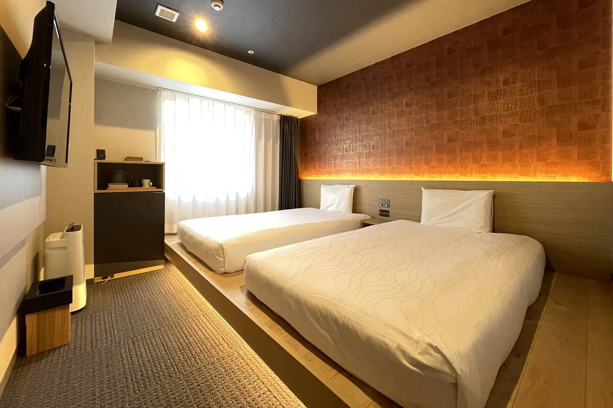 Une chambre d’hôtel avec deux lits, un téléviseur à écran plat, un bureau avec une chaise et une fenêtre avec des rideaux.