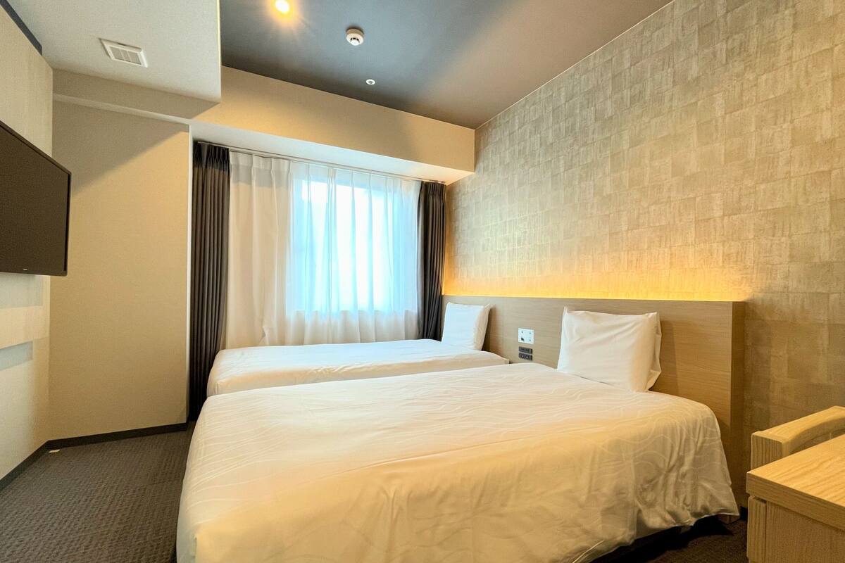 Une chambre d’hôtel avec deux lits, un téléviseur à écran plat, une fenêtre avec des rideaux et un mur à texture.