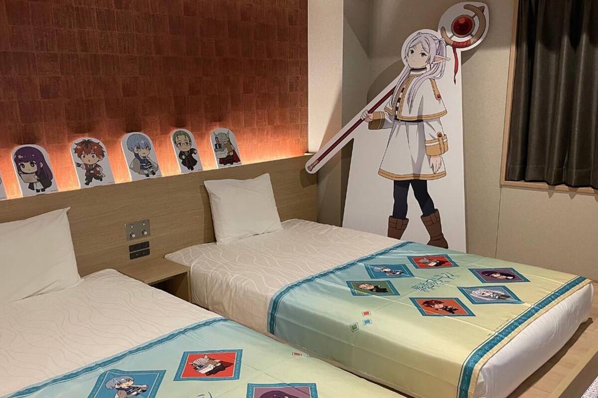 Une chambre d’hôtel avec deux lits, un mur en briques, des affiches de personnages d’anime et une grande découpe de personnage d’anime accrochée au mur.