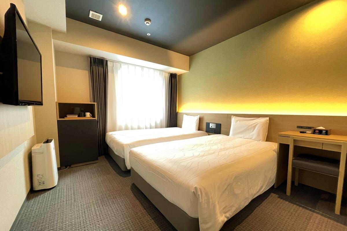 Une chambre d’hôtel avec deux lits, un téléviseur fixé au mur, une table de chevet avec une lampe et une petite étagère avec quelques objets.