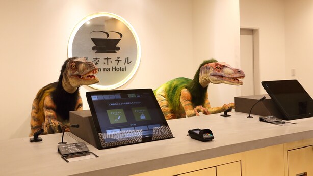 A reception desk with a digital display, a sign with a dinosaur illustration, and a camera on the ceiling.