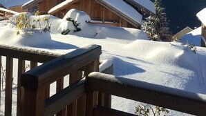 Un chalet de madera con techo cubierto de nieve, frente a montañas coronadas por nieve bajo un cielo azul despejado.