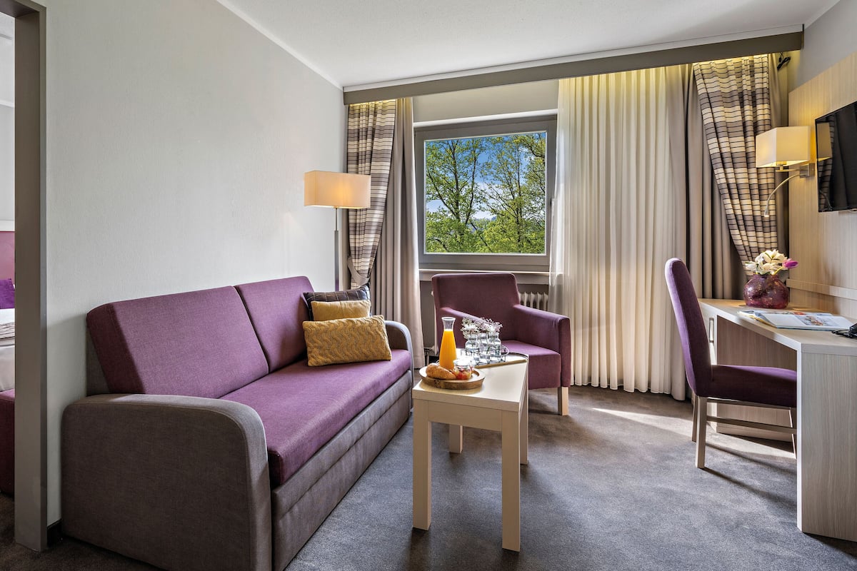 Ein Hotelzimmer mit violettem Sofa, einem Bett mit weißer Bettwäsche, einem kleinen Tisch mit Vase und Tablett sowie einem Schreibtisch mit Stuhl.
