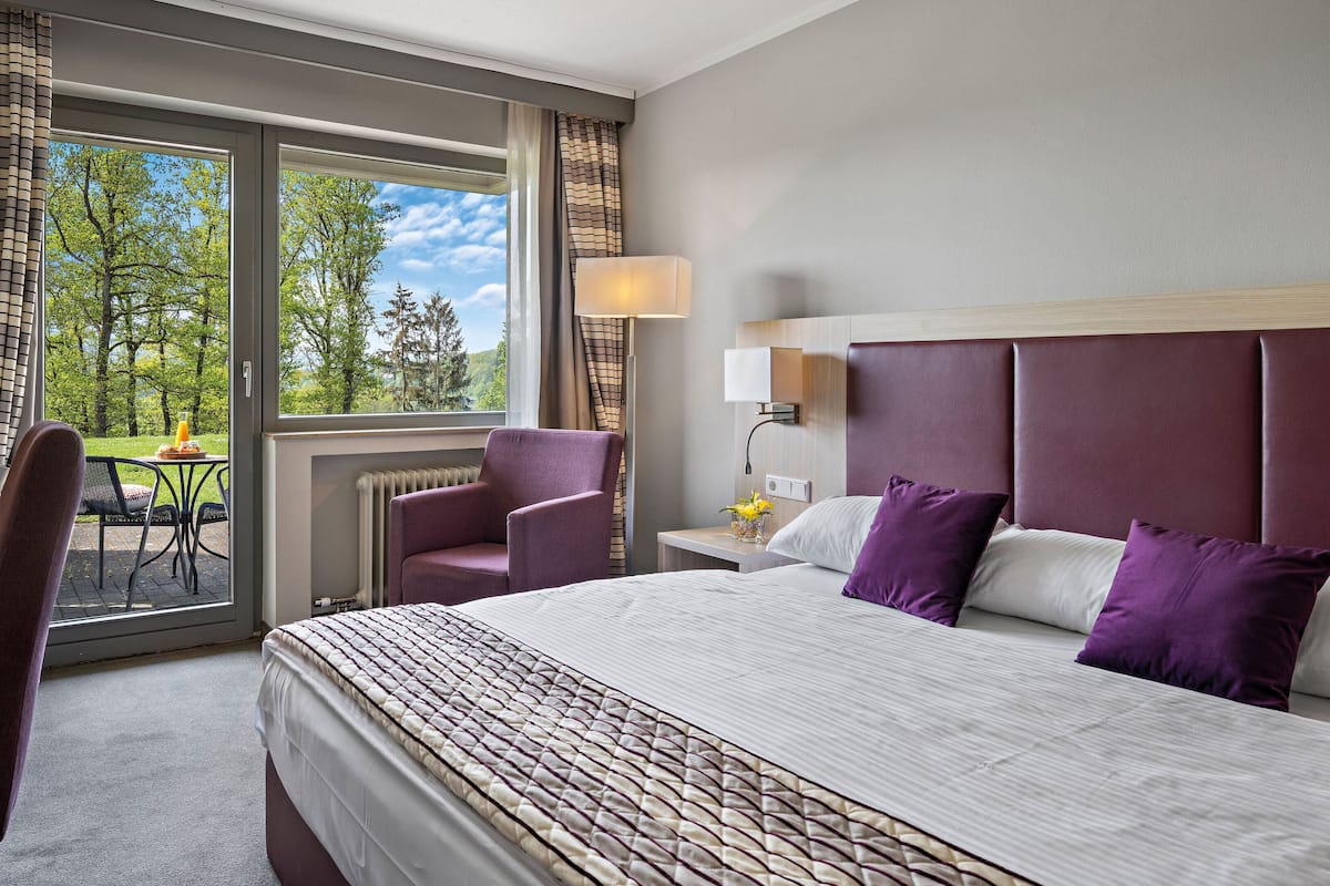 Ein Hotelzimmer mit einem großen Bett, einem Schreibtisch mit Stuhl, einem Tisch mit Stuhl und Blick auf Bäume durch das Fenster.