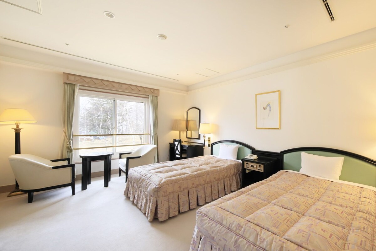 A hotel room with two beds, a desk, a chair, a window with curtains, and a wall-mounted lamp.