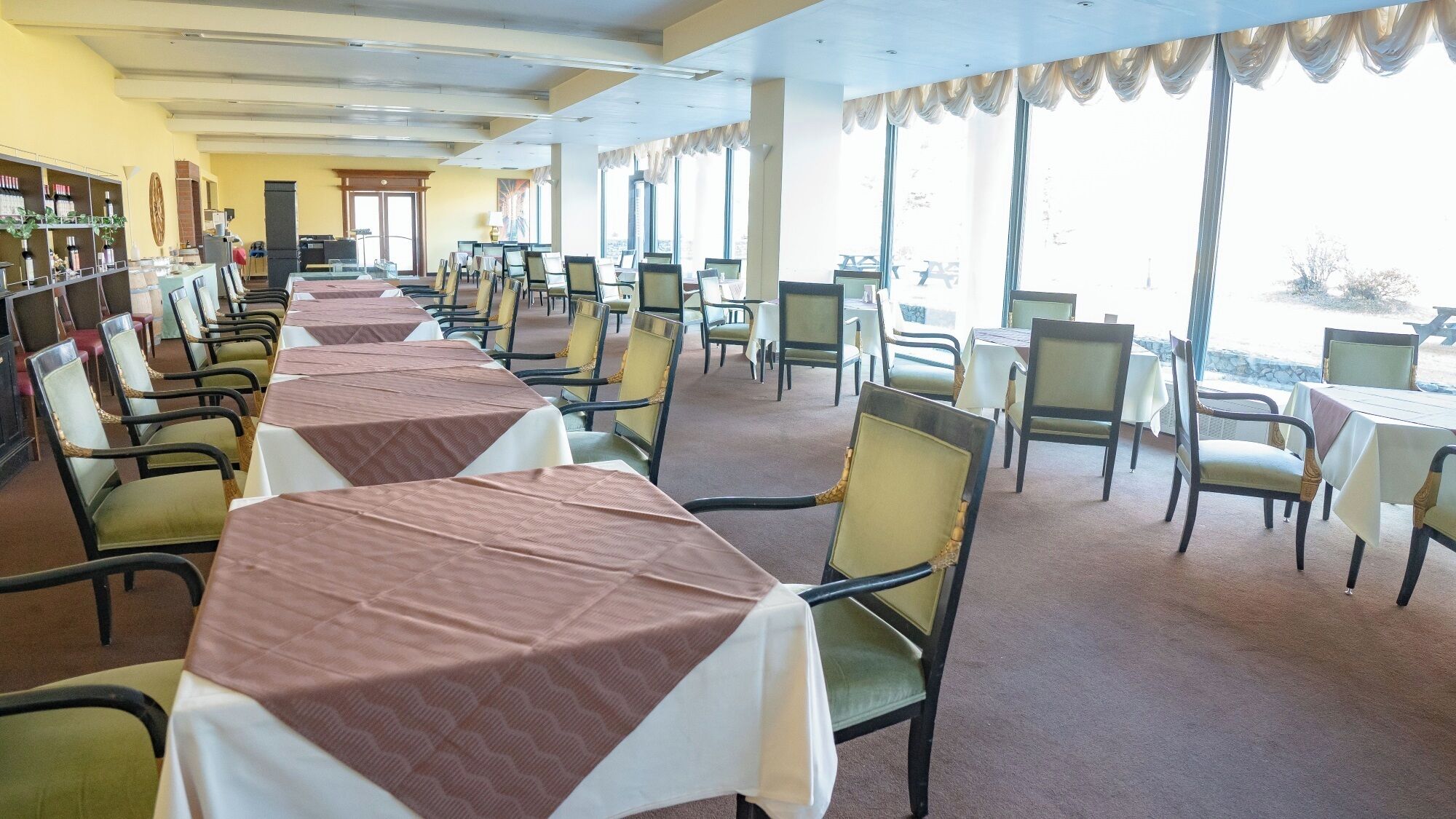 A restaurant with long tables and chairs, a bar area with bottles, and large windows offering a view.
