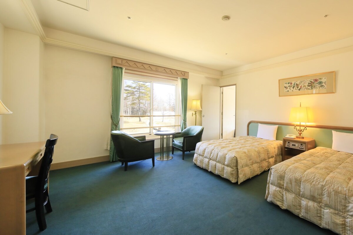 Une chambre d’hôtel avec deux lits, un bureau, une chaise et une fenêtre avec des rideaux.