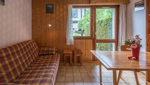 A cozy room with a plaid sofa, a wooden table, and a window with a view of greenery outside. - Cozy studio in Chamonix - Pets allowed, private parking (Chamonix-Mont-Blanc)