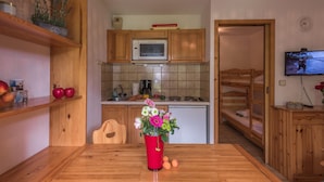 A compact kitchen with wooden cabinets, a microwave, and a sink. There is a dining table with a red vase of flowers and apples. A television is mounted on the wall, and a bunk bed is visible in the background. - Cozy studio in Chamonix - Pets allowed, private parking (Chamonix-Mont-Blanc)