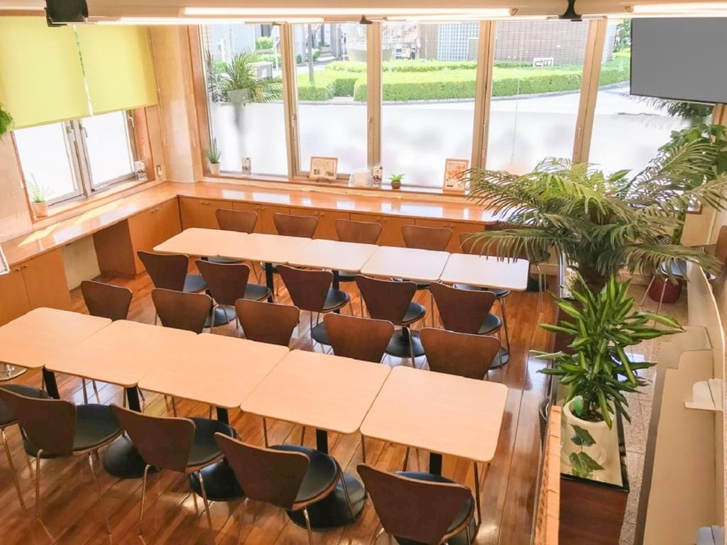 A conference room with a long table and chairs, large windows with a view of the street, and indoor plants.