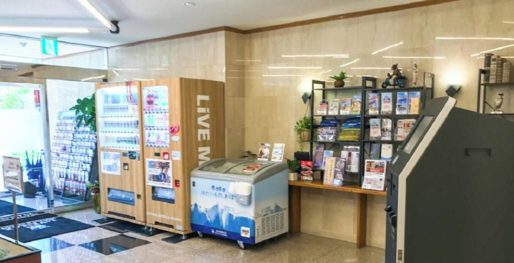Zona de máquinas expendedoras con un rótulo que dice 'LIVE', un refrigerador y un expositor de revistas.