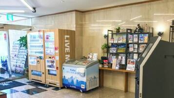 Zona de máquinas expendedoras con un rótulo que dice 'LIVE', un refrigerador y un expositor de revistas.