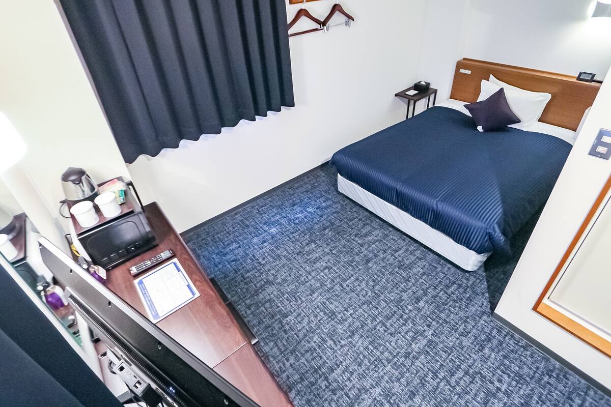 Quarto de hotel com cama, escrivaninha com computador, mesinha de apoio com abajur e janela com cortinas.
