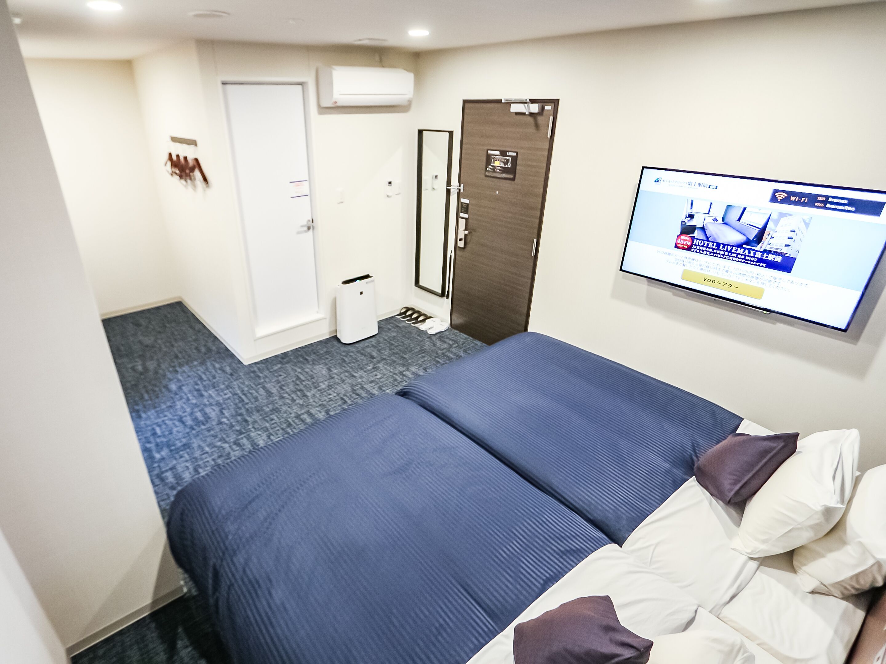 A hotel room with a bed, a TV, a door, and a small trash bin.