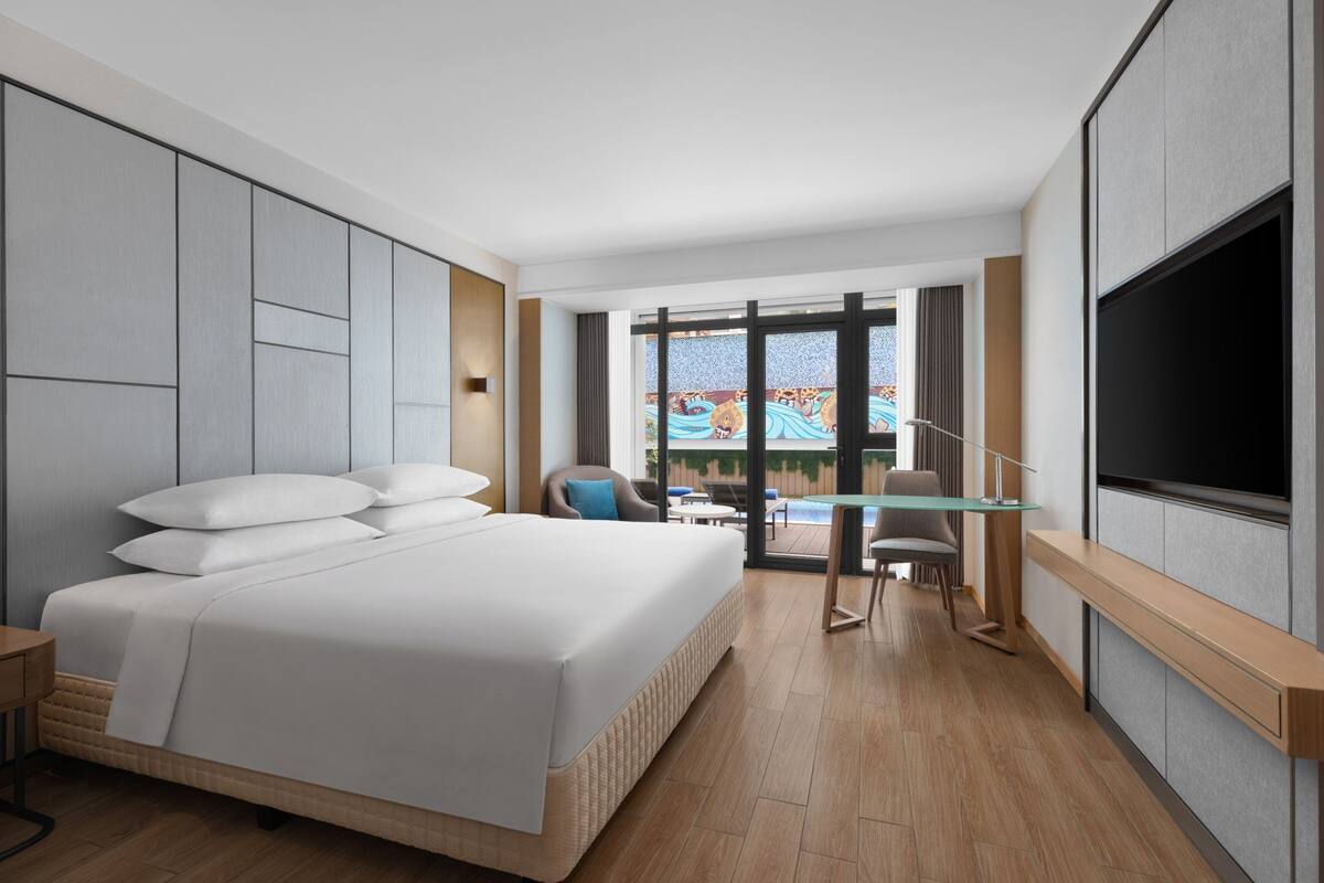 Habitación de hotel moderna con una cama grande, un televisor de pantalla plana, una mesa de vidrio y vistas a una piscina y zona exterior.