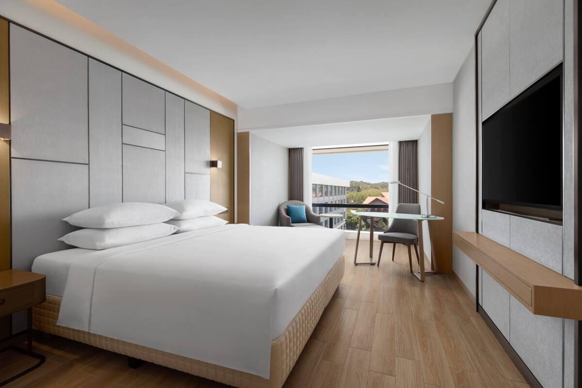 Una habitación de hotel moderna con una cama grande, un escritorio con silla, un televisor y un balcón con vistas.
