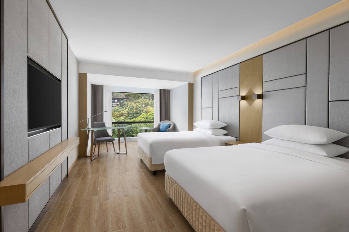 Habitación de hotel con dos camas, televisión, escritorio y balcón con vistas.