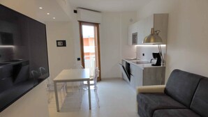 Een moderne woonkamer met een grijze bank, een eettafel met stoelen, een keuken met een spoelbak en een hanglamp, en een tv die aan de muur is bevestigd.