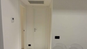 Een gang met een gesloten deur aan het einde, een lichtschakelaar en een klein ventilatierooster op de muur.