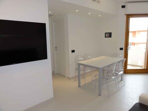 Un espace repas moderne avec une table blanche et des chaises transparentes, un téléviseur noir fixé au mur et une porte donnant sur un espace extérieur.
