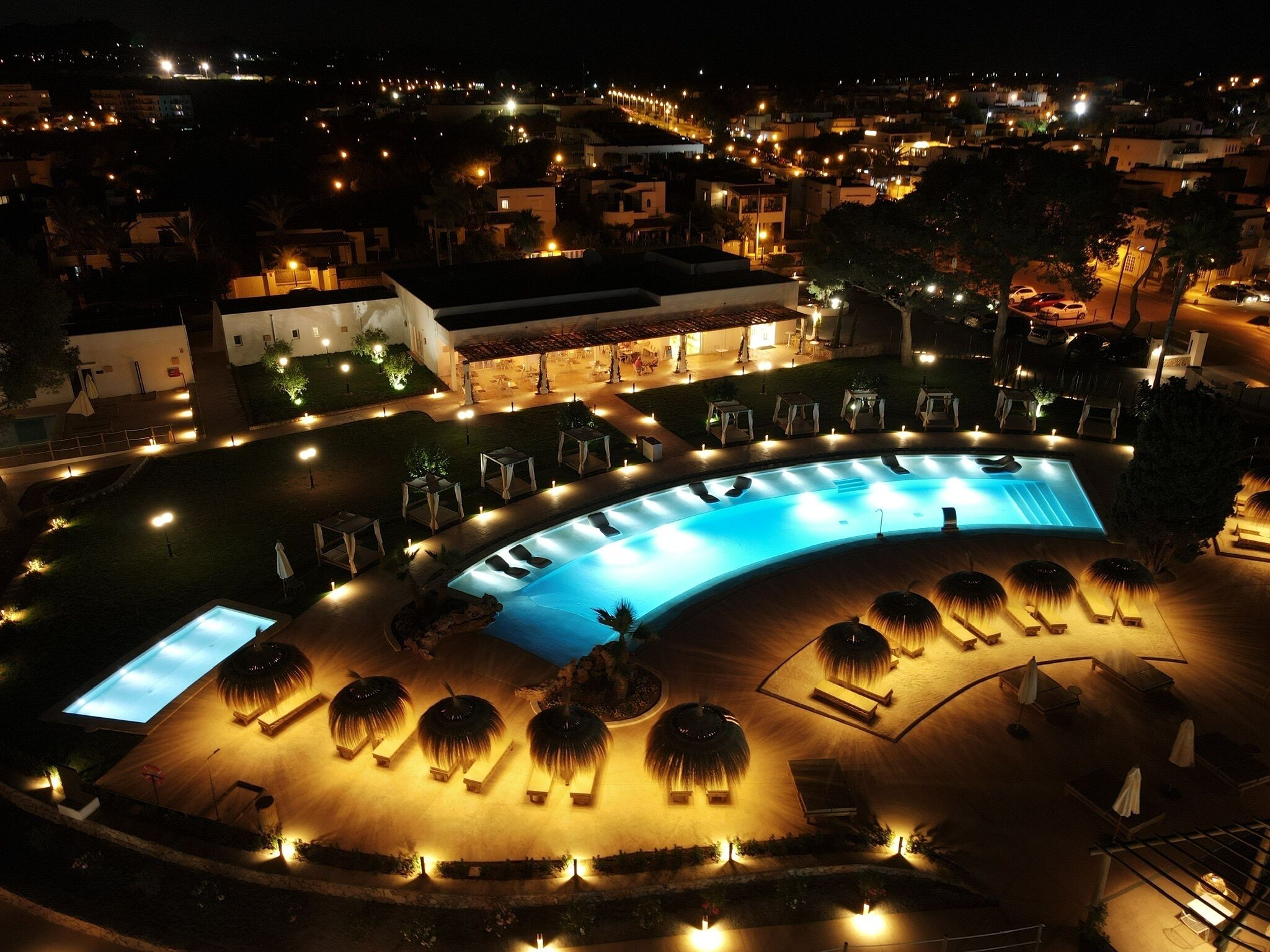 Vue nocturne d’un complexe hôtelier avec une grande piscine illuminée, des chaises longues et un espace extérieur bien éclairé.