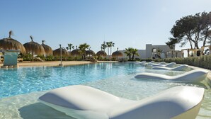 Zona de piscina con sillones de descanso, palmeras y sombrillas de paja.