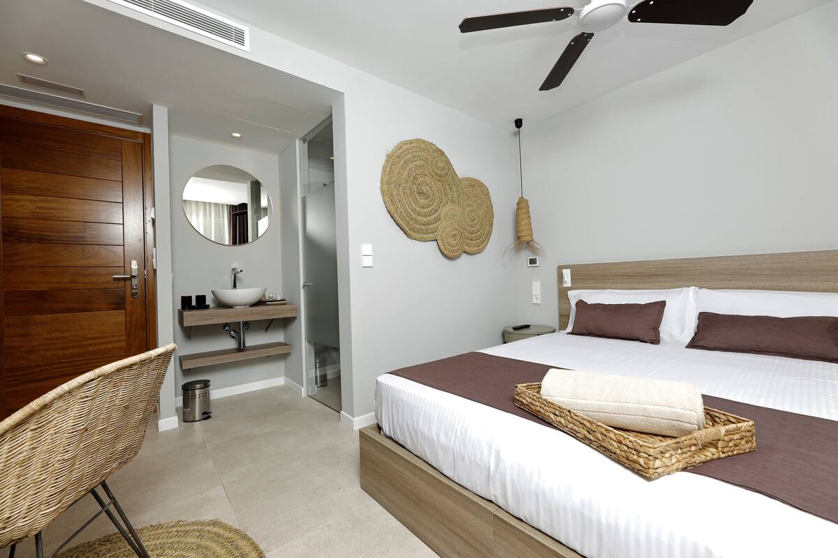 A modern bedroom with a wooden door, a round mirror, a large woven wall art piece, a ceiling fan, and a neatly made bed with a brown throw blanket.