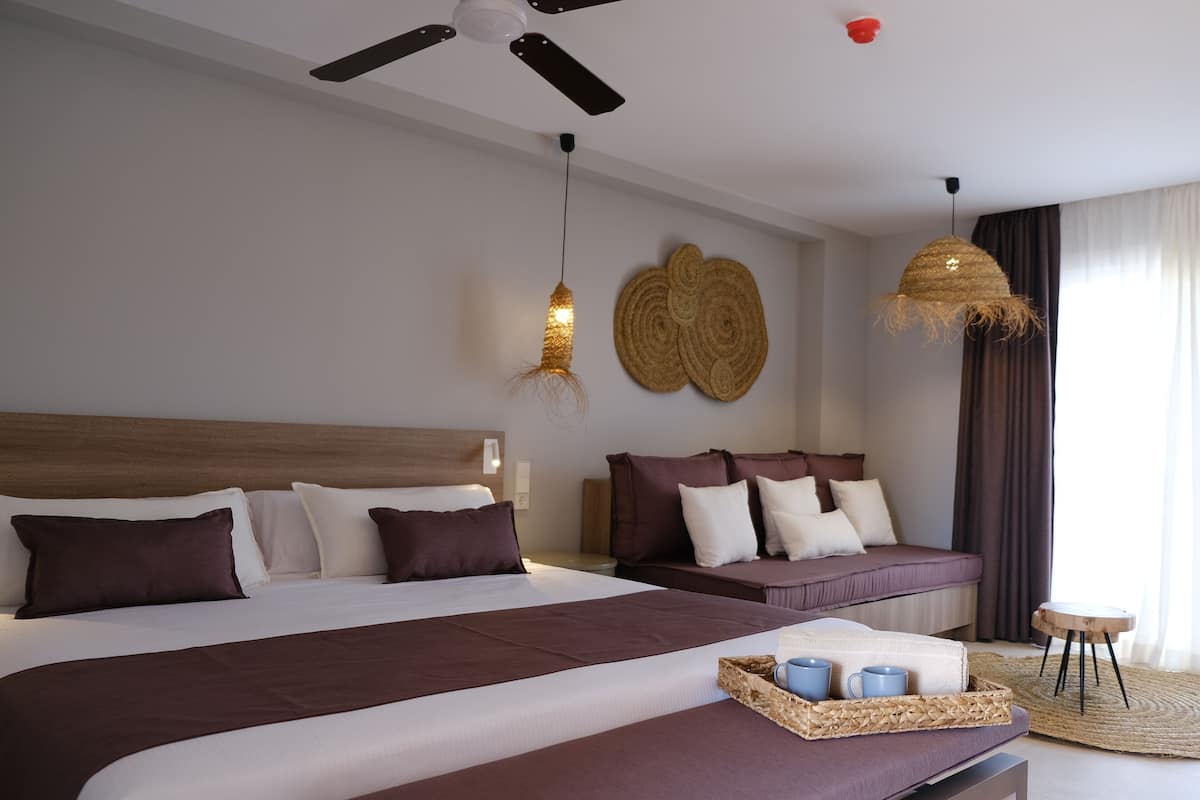 A modern hotel room with a large bed, a sofa, a ceiling fan, and a woven light fixture.