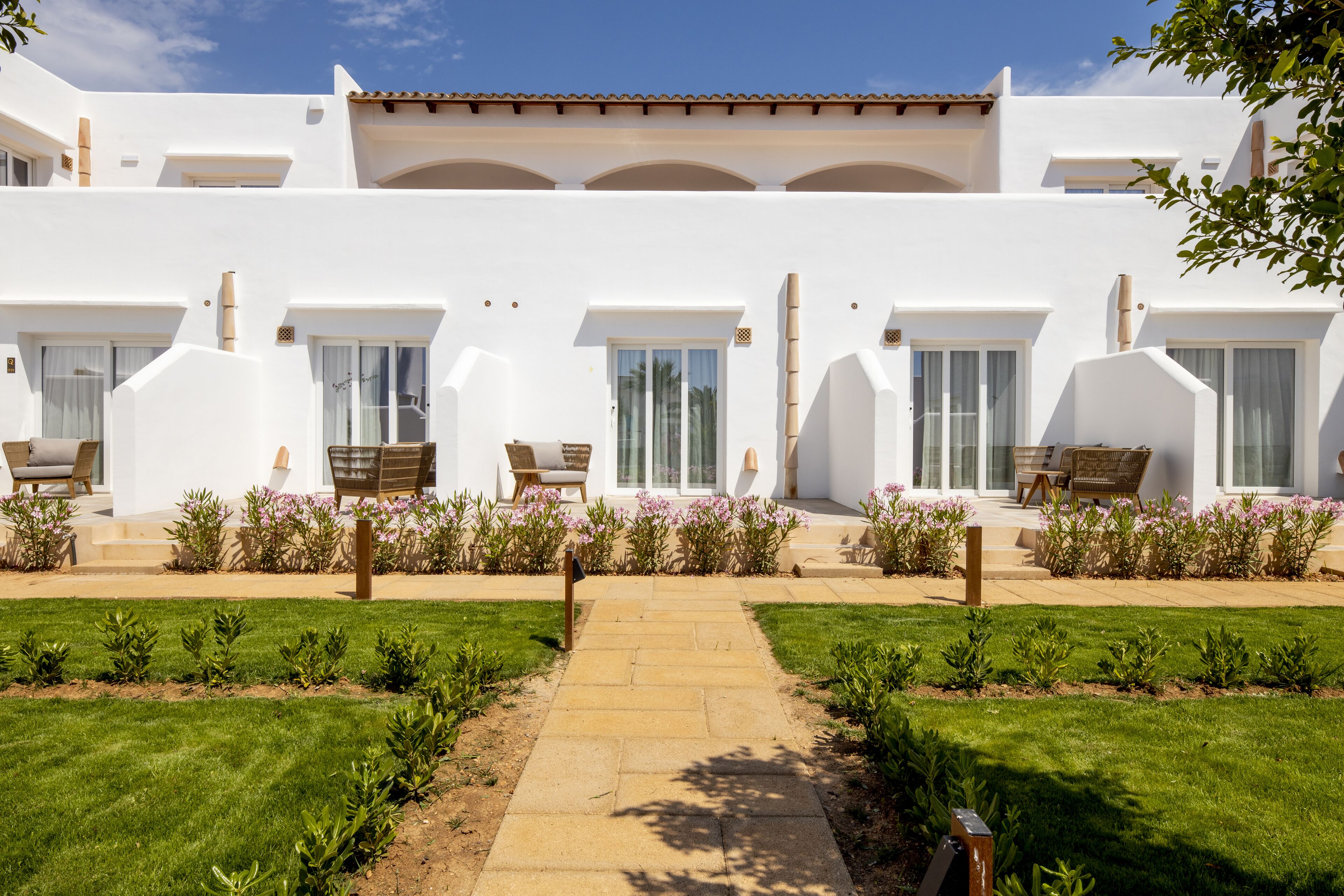A white building with multiple balconies and large windows, surrounded by a well-maintained garden with a stone pathway leading to the entrance.