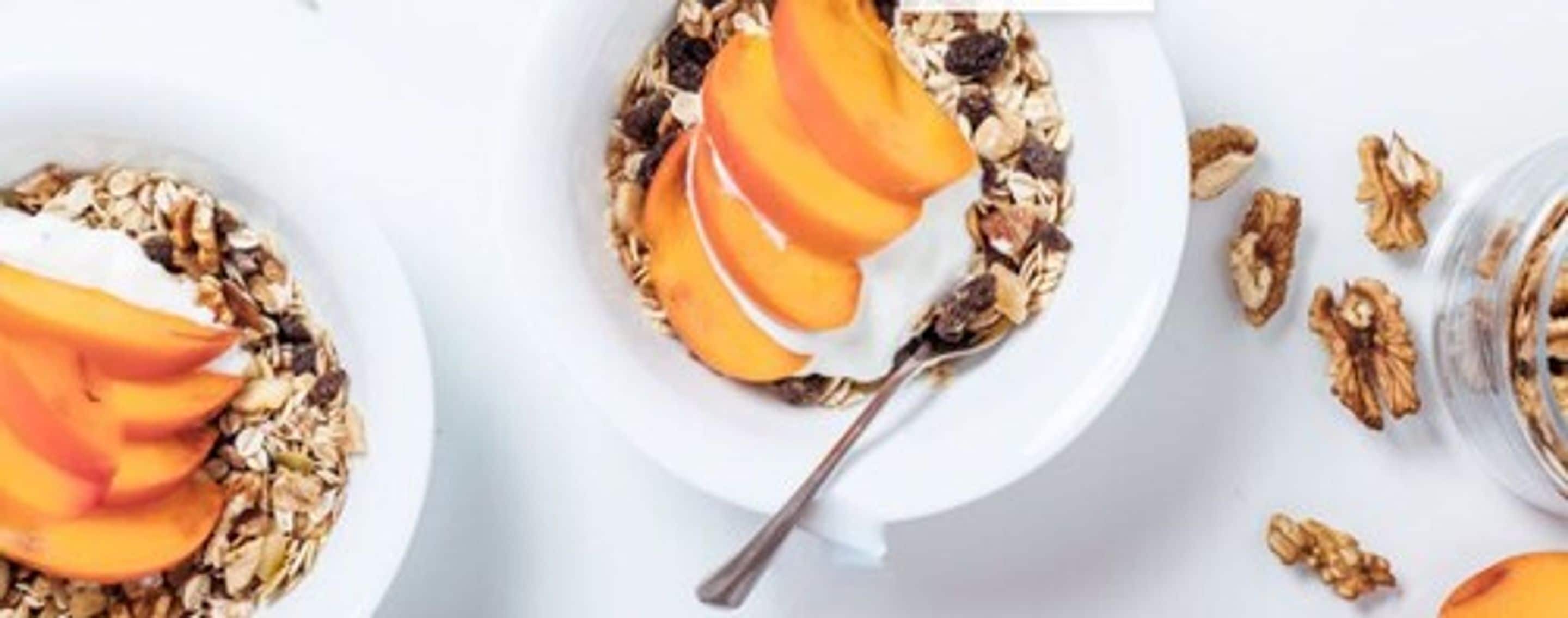 Colazione con granola, pesche a fettine e noci su un piatto bianco.