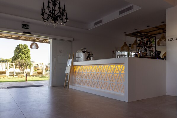 Un espace bar moderne avec un comptoir blanc, un lustre et une vue sur l’extérieur.